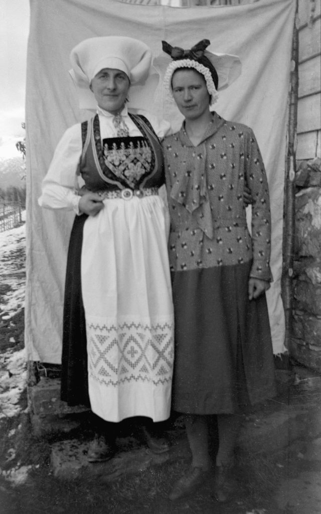Portrait of unknown women in traditional dress The photo is probably taken at their house. The woman to the left is wearing the bunad and headdress from Hardager, the woman to the right is in modern dress, with the traditional headdress from Sogn. ID: EL.0045.0005 Photographer: Elen Loftesnes